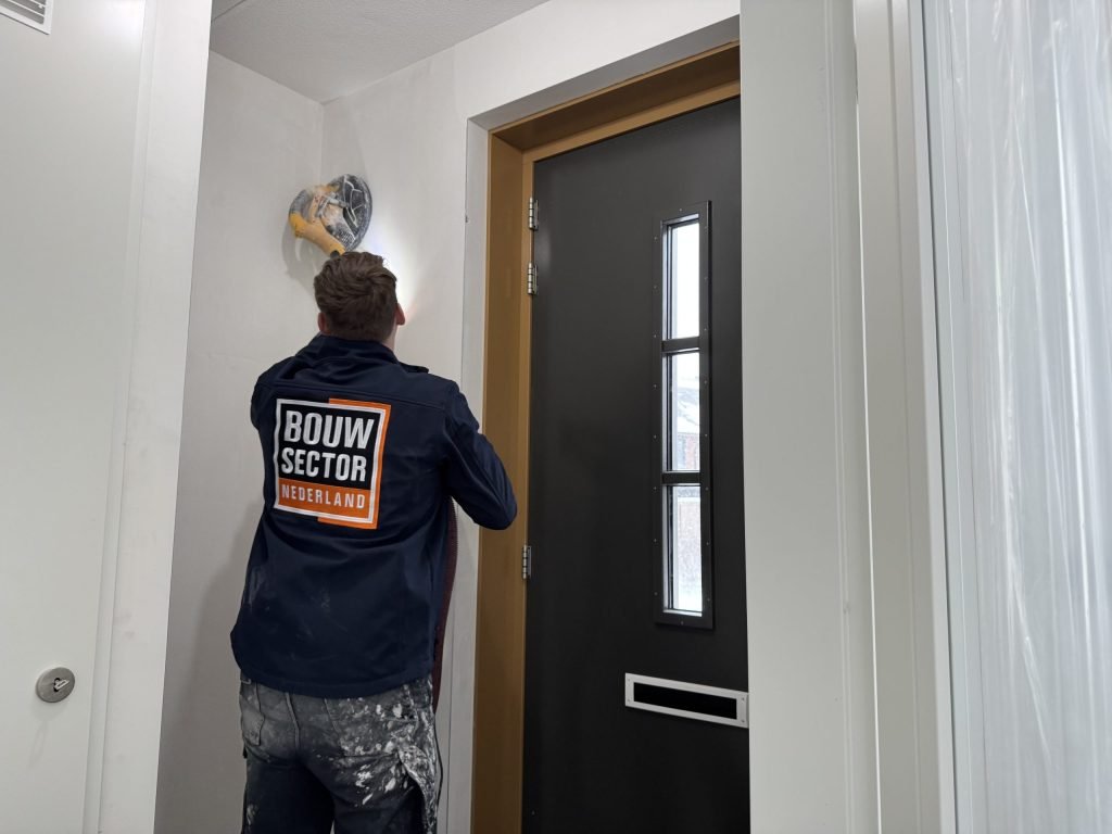 Vakman van Bouwsector Nederland schuurt boven een voordeur met een wandschuurmachine met afzuiging