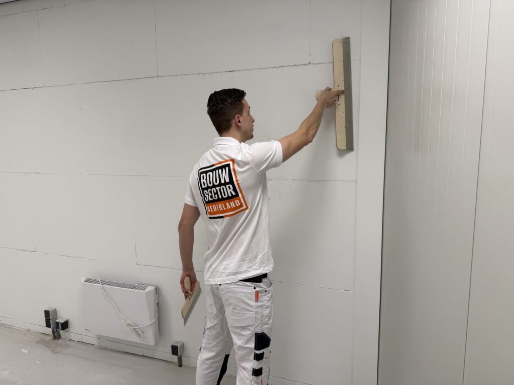 Stukadoor van Bouwsector Nederland zet een lange spaan hoog tegen een wand, met een tweede spaan in zijn andere hand