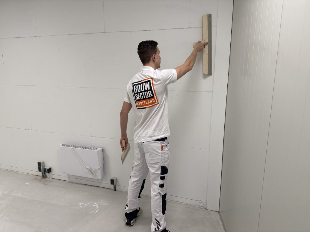 Stukadoor in wit werkpak zet een spaan tegen een wand in een lege ruimte met een elektrische kachel
