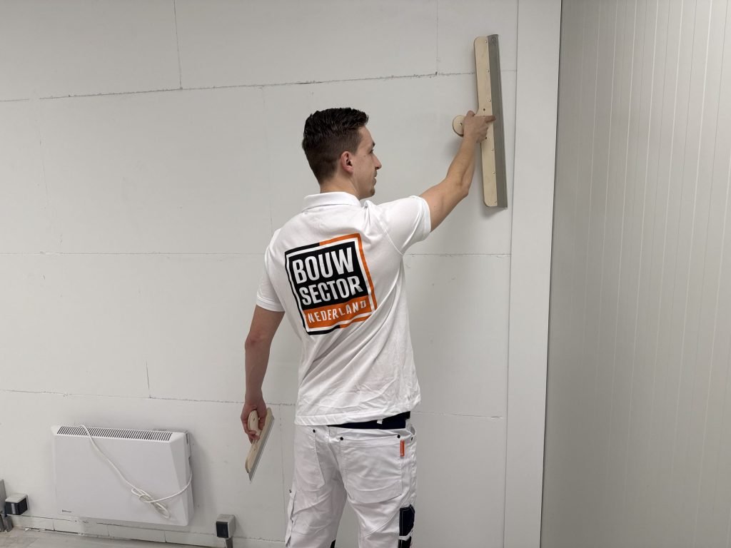 Stukadoor in een wit shirt met het logo van Bouwsector Nederland trekt een spaan over een lichte wand