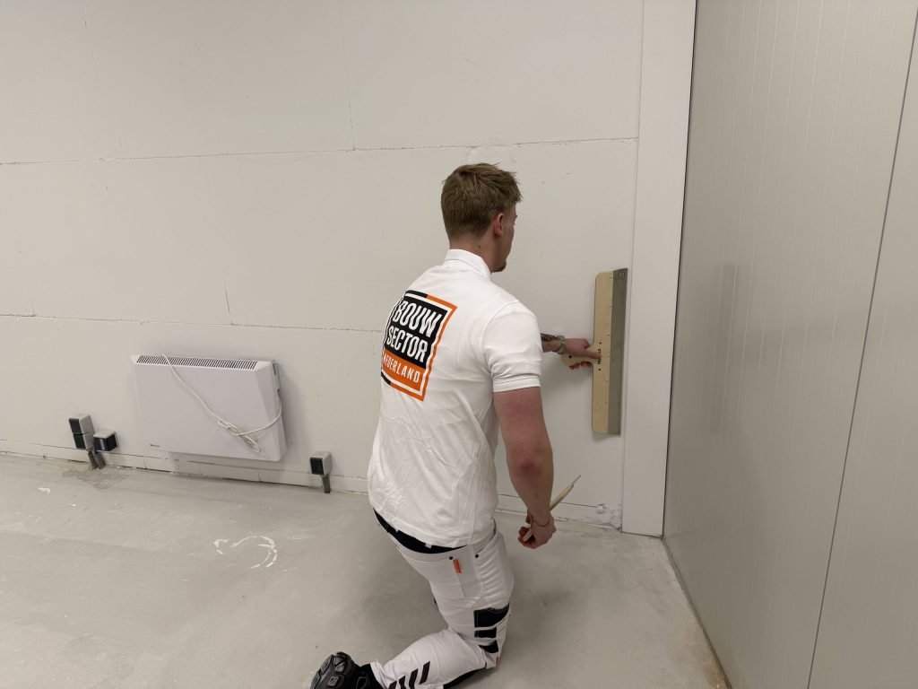 Stukadoor van Bouwsector Nederland zit op een knie en houdt een lange spaan onderaan tegen de wand