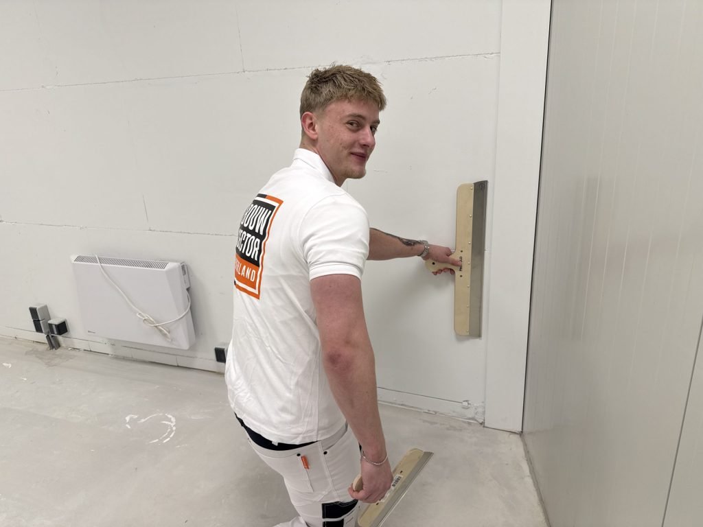 Lachende stukadoor van Bouwsector Nederland met een spaan tegen de wand en een tweede spaan in de hand