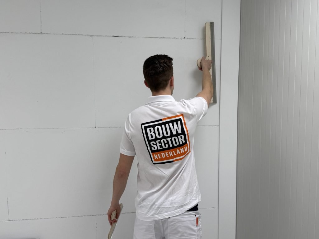 Stukadoor met het logo van Bouwsector Nederland op zijn rug trekt een lange spaan verticaal langs een wand
